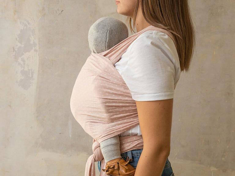 Frau im Profil trägt ein Baby in einem rosa Tragetuch vor der Brust, das Baby trägt eine graue Strickmütze und braune Schuhe