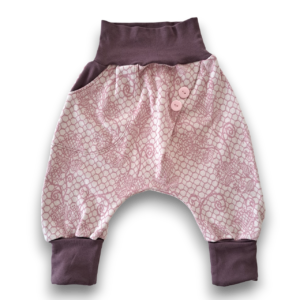 Locker geschnittene Kinderhose aus cremefarbenem Baumwolljacquard mit zartrosa Rankenmuster und zwei rosa Knöpfen am Bund, Bündchen in dunklem Mauve.