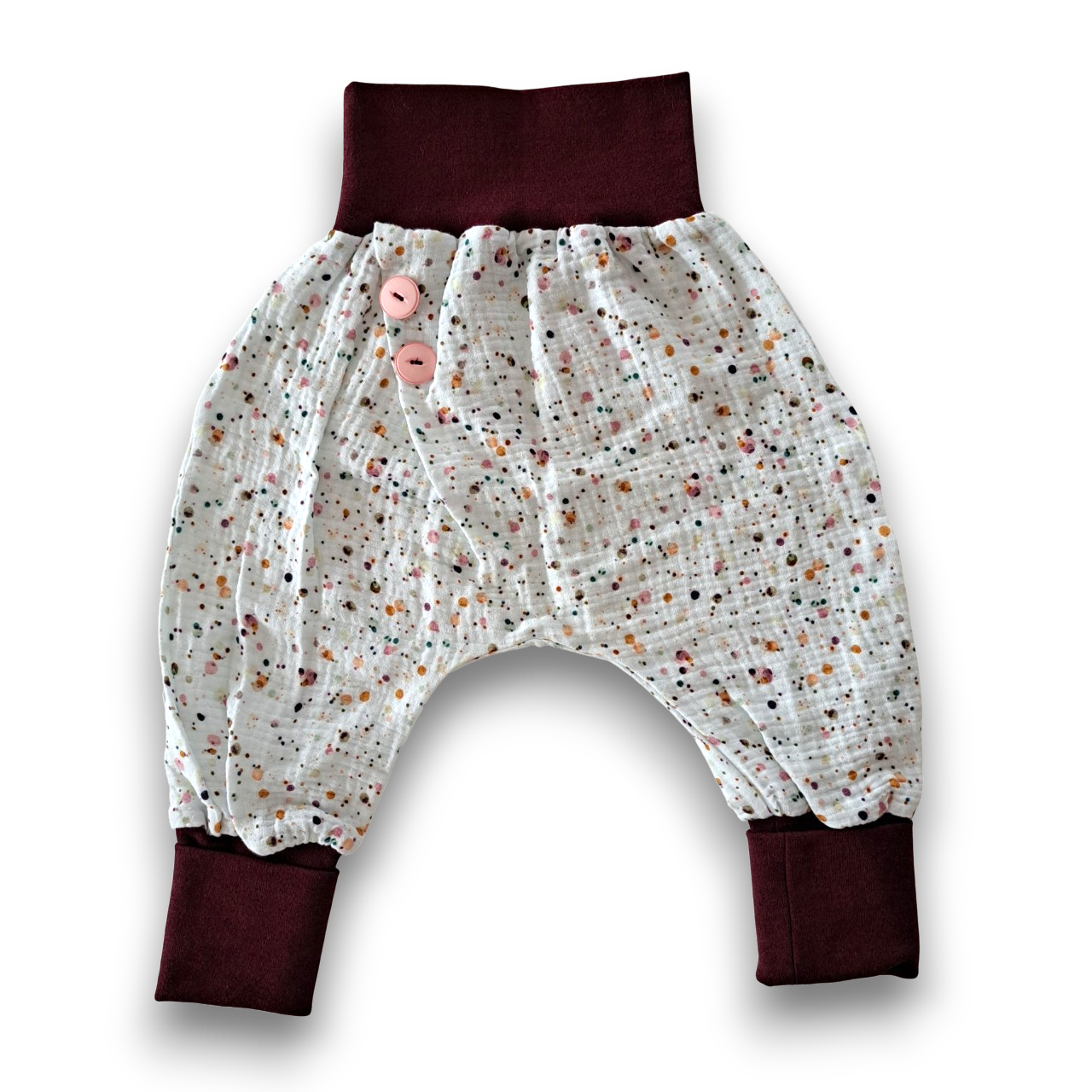 Lockere Kinderhose aus cremefarbenem Musselin mit buntem Konfetti-Muster, dunkelroten Bündchen und zwei rosa Knöpfen.