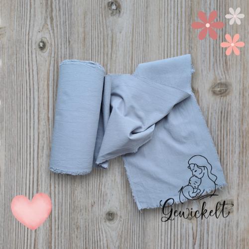 Gerollter grauer Bengkung-Belly-Binding-Tuch aus Stoff, mit einem naturfarbenen Band zusammengebunden, auf hellem Holzuntergrund, dekoriert mit Herz- und Blumen-Grafiken sowie dem Logo „Gewickelt“