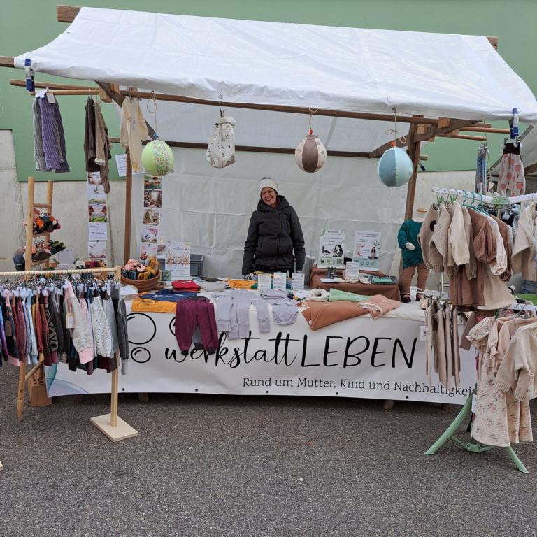 Marktstand von „werkstattLEBEN“ mit nachhaltig hergestellter Kleidung und Accessoires für Mutter und Kind: Im Vordergrund Kleiderständer mit bunter Baby- und Kinderkleidung, auf dem Tisch Bodyes und Textilien, rechts Strickjacken und Hosen, im Hintergrund eine Person am Stand unter einem weissn Zeltdach.